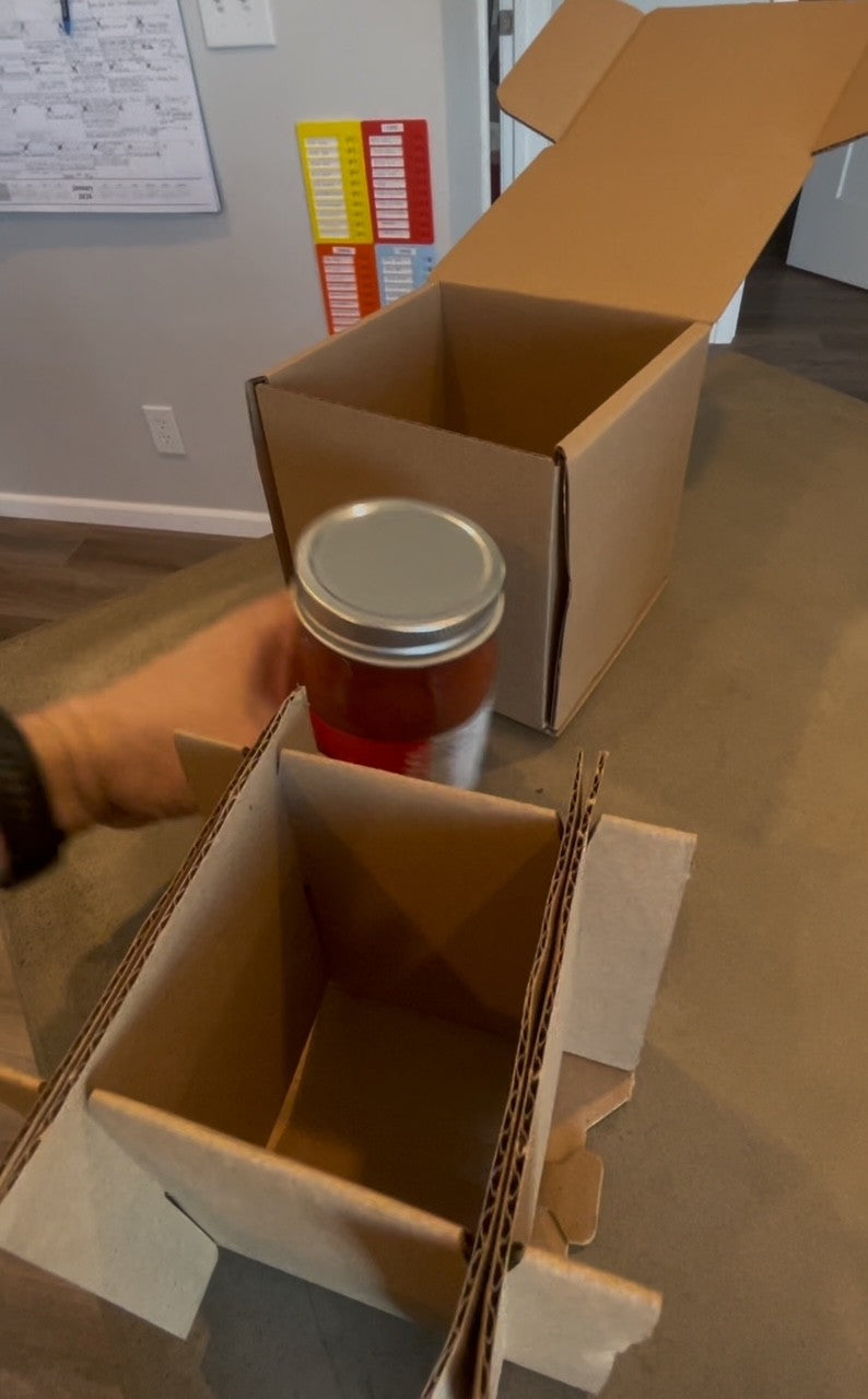 Person holding a jar over an open cardboard box on a wooden floor.

